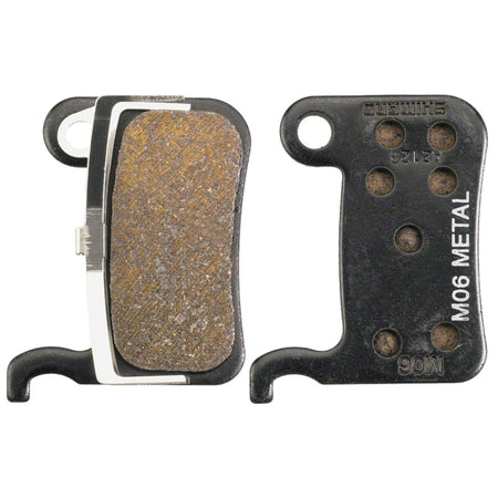 Shimano M06 metal disc brake pads for bicycles, showing the brake pads with textured metal surface and mounting hardware.